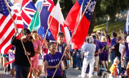 一群欢笑的学生举着不同国家的国旗参加TCU返校节游行, 由一个穿着印有“青蛙”字样衬衫的女孩带领